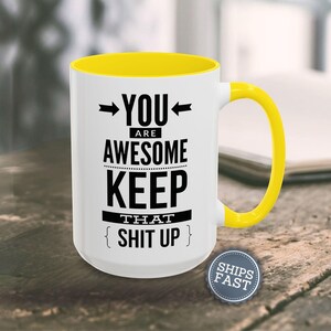 A white coffee mug with a yellow handle, displaying a humorous text that reads "YOU ARE AWESOME KEEP THAT SHIT UP" along with a small "SHIES FAST" logo.