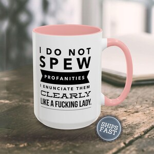 A white mug with a pink handle, featuring a humorous text that reads "I DO NOT SPEW PROFANITIES I ENUCIATE THEM CLEARLY LIKE A FUCKING LADY."