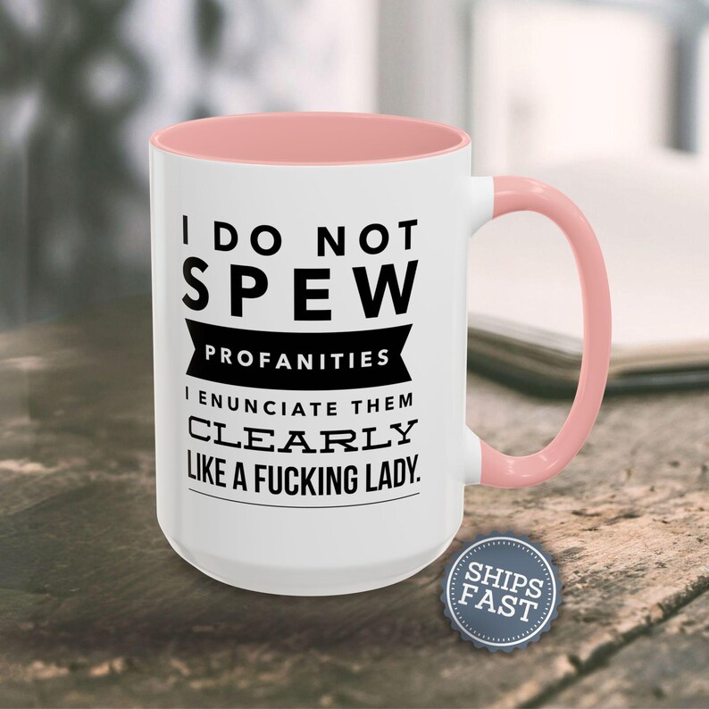 A white mug with a pink handle, featuring a humorous text that reads "I DO NOT SPEW PROFANITIES I ENUCIATE THEM CLEARLY LIKE A FUCKING LADY."