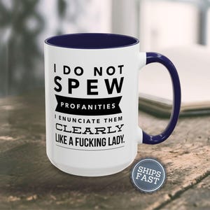 A white mug with a blue handle, featuring a humorous text that reads "I DO NOT SPEW PROFANITIES I ENUCATE THEM CLEARLY LIKE A FUCKING LADY."