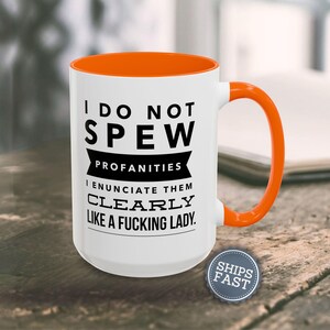 A white coffee mug with an orange handle, displaying a humorous text that reads "I DO NOT SPEW PROFANITIES I ENUCIATE THEM CLEARLY LIKE A FUCKING LADY."
