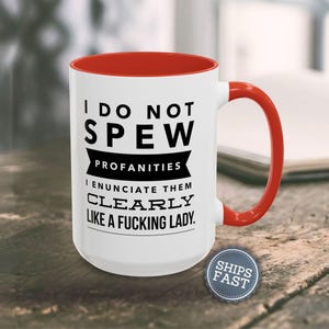 A white mug with a red handle, featuring a humorous text that reads "I DO NOT SPEW PROFANITIES I ENUCIATE THEM CLEARLY LIKE A FUCKING LADY."
