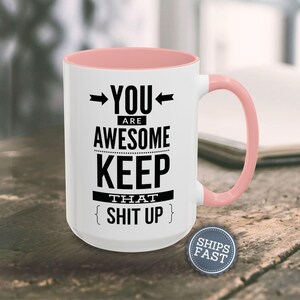 A white mug with a pink handle, displaying a humorous text that reads "YOU ARE AWESOME KEEP THAT SHIT UP" along with a small "SHIES FAST" logo.