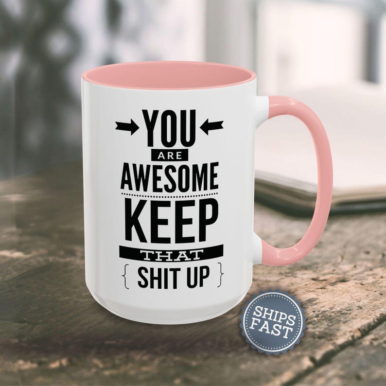 A white mug with a pink handle, displaying a humorous text that reads "YOU ARE AWESOME KEEP THAT SHIT UP" along with a small "SHIES FAST" logo.