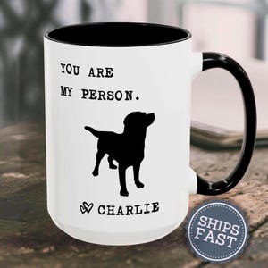 May include: A white ceramic mug with a black interior and handle. The front features the black text "YOU ARE MY PERSON." above a black silhouette of a dog, followed by two small heart icons and the name "CHARLIE" in black capital letters.