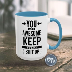 A white and blue mug with the text "YOU ARE AWESOME KEEP THAT SHIT UP" printed on it, along with a blue handle.