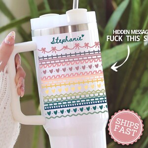 May include: A white stainless steel tumbler with a handle and straw, featuring the name Stephanie in teal script, followed by rows of colorful bows, hearts, and geometric patterns. The text HIDDEN MESSAGE FUCK THIS SHIT is visible.