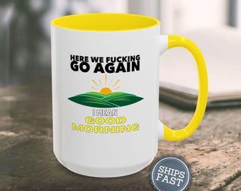 Pode incluir: Caneca de cerâmica branca com interior e alça amarelos. A caneca apresenta o texto "HERE WE FUCKING GO AGAIN" acima de um gráfico de um nascer do sol sobre colinas verdes, com "I MEAN GOOD MORNING" abaixo. Um adesivo "SHIPS FAST" é visível.