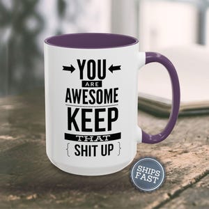 A white mug with a purple handle, displaying a humorous text that reads "YOU ARE AWESOME KEEP THAT SHIT UP" along with a small "SHIES FAST" logo.