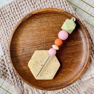 Peut inclure: Une assiette en bois avec un biscuit hexagonal et une brochette en métal avec des perles de couleur pastel. Les perles sont vertes, roses, orange et roses.