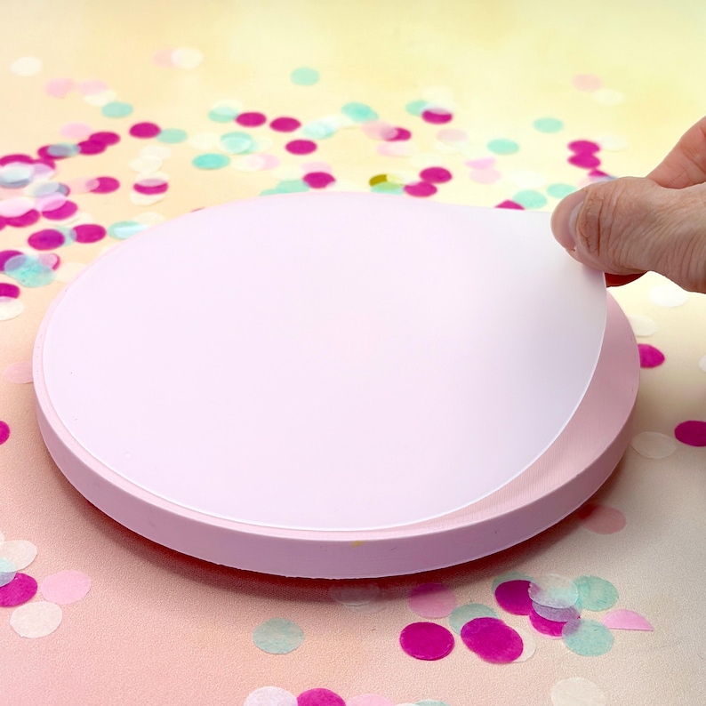 Pode incluir: Um prato girat&oacute;rio redondo rosa para bolos com um c&iacute;rculo de papel branco no topo. O c&iacute;rculo de papel est&aacute; sendo levantado por uma m&atilde;o.
