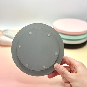 Pode incluir: Um coaster redondo cinza com uma superf&iacute;cie lisa e seis pequenos p&eacute;s de borracha na parte inferior.