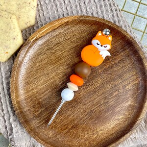 Könnte beinhalten: Ein Holzteller mit einer Silikon-Fuchsperle und bunten Perlen auf einem silbernen Spieß. Die Perlen sind braun, orange und weiß.