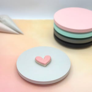 Pode incluir: Um biscoito em forma de cora&ccedil;&atilde;o rosa em um suporte de bolo branco. O suporte est&aacute; em um fundo laranja claro.