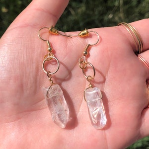 May include: A pair of gold earrings with clear quartz crystal pendants. The earrings have a simple design with a small gold hoop and a delicate wire wrapped around the crystal.