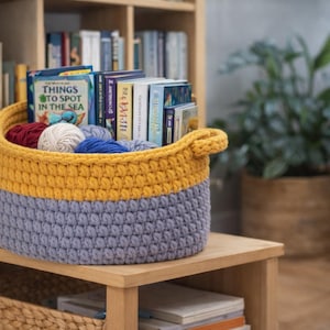 May include: A handmade crochet basket in yellow and gray, filled with books and yarn. The basket has a sturdy, textured appearance and is placed on a wooden shelf. The books include titles like "Things to Spot in the Sea."