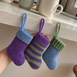 May include: Three small, hand-knitted stockings with hanging loops. The first is purple with a light blue cuff. The second features purple and green horizontal stripes with a purple cuff. The third is light blue with a green ribbed cuff.