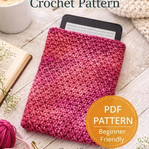 May include: A pink and magenta crochet Kindle sleeve with a Kindle inside. The image includes the text "Kindle Sleeve Crochet Pattern" and a "PDF Pattern Beginner Friendly" sticker. Yarn and a crochet hook are also visible.