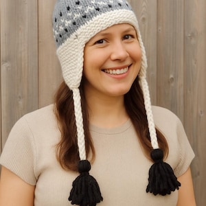 Pode incluir: Um gorro de malha com abas para as orelhas, um pompom preto e borlas. O chapéu é cinzento com padrões geométricos brancos e pretos. O texto "PDF PATTERN" é exibido na parte inferior da imagem.