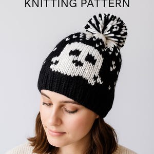 Może przedstawiać: Czarno-biała dzianinowa czapka z motywem ducha i pomponem. Na czapce widnieje napis "Ghost Hat Knitting Pattern". Czapkę nosi osoba w kremowym swetrze.