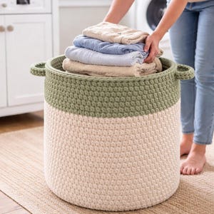 Può includere: Un grande cesto per la biancheria all'uncinetto a due toni. La sezione superiore è verde oliva e quella inferiore color crema. Il cesto ha due manici ed è pieno di asciugamani piegati nelle tonalità del beige e del blu.