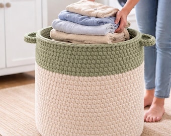May include: A large, two-toned crochet laundry basket. The top section is olive green, and the bottom is cream-colored. The basket has two handles and is filled with folded towels in shades of beige and blue.