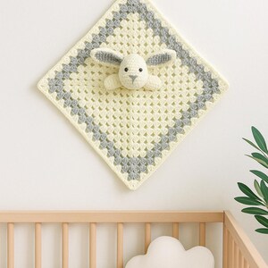 May include: A handmade, cream-colored crochet baby blanket with a gray border and a 3D bunny head. The blanket is square-shaped and hangs on a white wall. A wooden crib and cloud-shaped pillow are in the foreground.