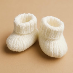 Puede incluir: Un par de patucos de bebé de punto de color crema. Los patucos tienen un puño acanalado y un aspecto suave y texturizado. El fondo es de color beige neutro.