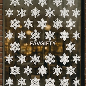 Puede incluir: Una ventana de vidrio decorada con numerosas calcomanías de copos de nieve blancos. La palabra "FAVGIFTY" se muestra en el centro. La ventana está enmarcada por un borde negro y colocada contra una pared de ladrillos.