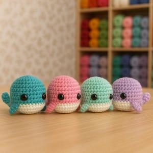 May include: Four colorful, crocheted whale figurines in a row. Each whale is a different color: teal, pink, mint green, and purple. They have white bellies and black button eyes. The whales are sitting on a light brown surface.
