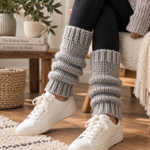 May include: A pair of light gray chunky knit leg warmers featuring a ribbed texture, worn over black leggings and paired with white lace-up sneakers. The leg warmers extend from the lower calf to the ankle, showcasing a thick, soft yarn construction.
