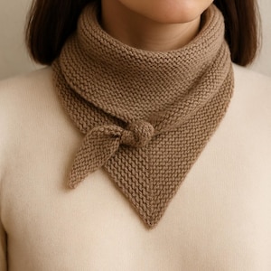 Peut inclure: Écharpe tricotée de couleur beige avec un nœud. L'écharpe est de forme triangulaire et semble être faite d'un fil doux et texturé. Elle est portée autour du cou et nouée sur le devant.