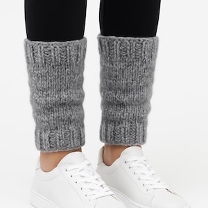 May include: Grey knitted leg warmers are worn over black leggings, paired with white trainers. The leg warmers have a chunky knit, offering a textured contrast to the smooth leggings and shoes. The trainers have white laces.