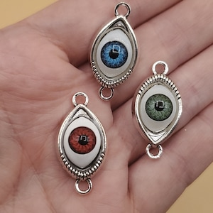 May include: Three silver eye charm pendants with different colored eyes. The eyes are blue, green, and brown. The charms are oval shaped and have a silver chain attached to the top.