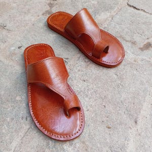 May include: A pair of brown leather sandals with a toe loop design. The sandals are hand-stitched with a light brown thread and have a simple, classic style. The sandals are shown on a stone surface.