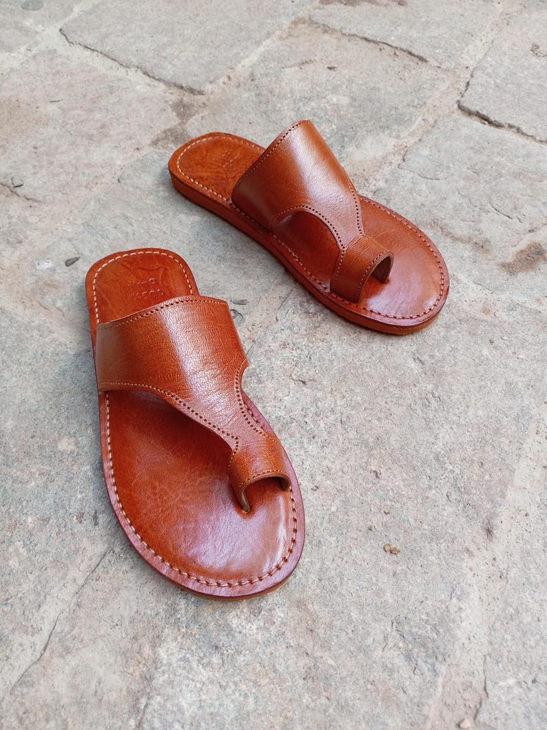 May include: A pair of brown leather sandals with a toe loop design. The sandals are hand-stitched with a light brown thread and have a simple, classic style. The sandals are shown on a stone surface.