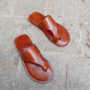 May include: A pair of brown leather sandals with a thong-style toe post and a strap across the top of the foot. The sandals are stitched around the edges and appear to be handmade. The sandals are displayed on a stone surface.