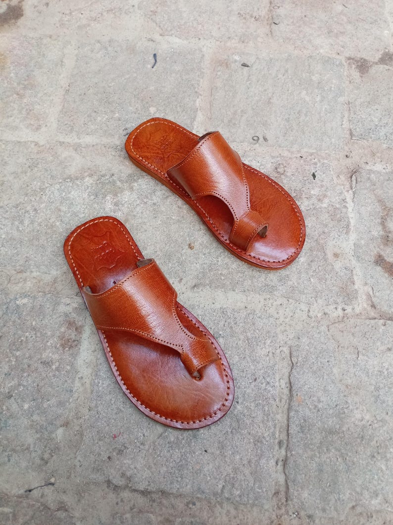 May include: A pair of brown leather sandals with a thong-style toe post and a strap across the top of the foot. The sandals are stitched around the edges and appear to be handmade. The sandals are displayed on a stone surface.