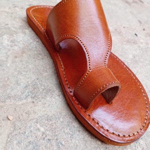 May include: A handcrafted, brown leather sandal with a toe loop and a wide strap. The sandal features detailed stitching around the edges and a smooth, polished finish. The footwear is designed for comfort and style.