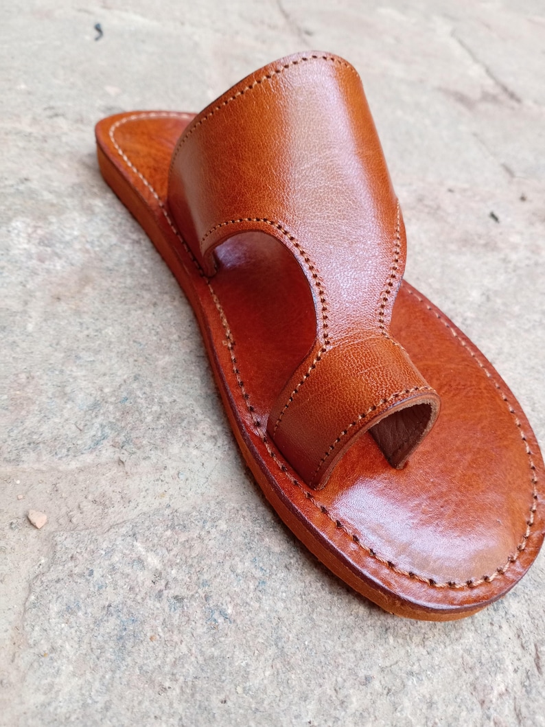 May include: A handcrafted, brown leather sandal with a toe loop and a wide strap. The sandal features detailed stitching around the edges and a smooth, polished finish. The footwear is designed for comfort and style.