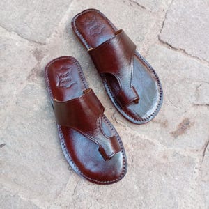 May include: A pair of brown leather sandals with a toe loop design. The sandals have a smooth, glossy finish and are stitched around the edges. The sandals are displayed on a stone surface.