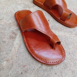 May include: A pair of brown leather sandals with a thong-style toe strap. The sandals have a stitched sole and a wide strap across the top of the foot. The leather has a smooth, polished finish.