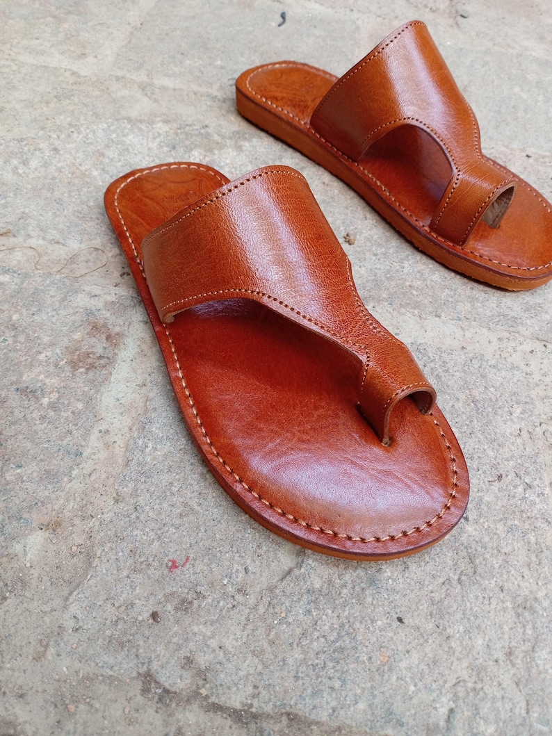 May include: A pair of brown leather sandals with a thong-style toe strap. The sandals have a stitched sole and a wide strap across the top of the foot. The leather has a smooth, polished finish.