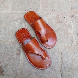 May include: A pair of brown leather sandals with a thong-style design. The sandals have a smooth, polished appearance and are stitched around the edges. The sandals are designed for casual wear.