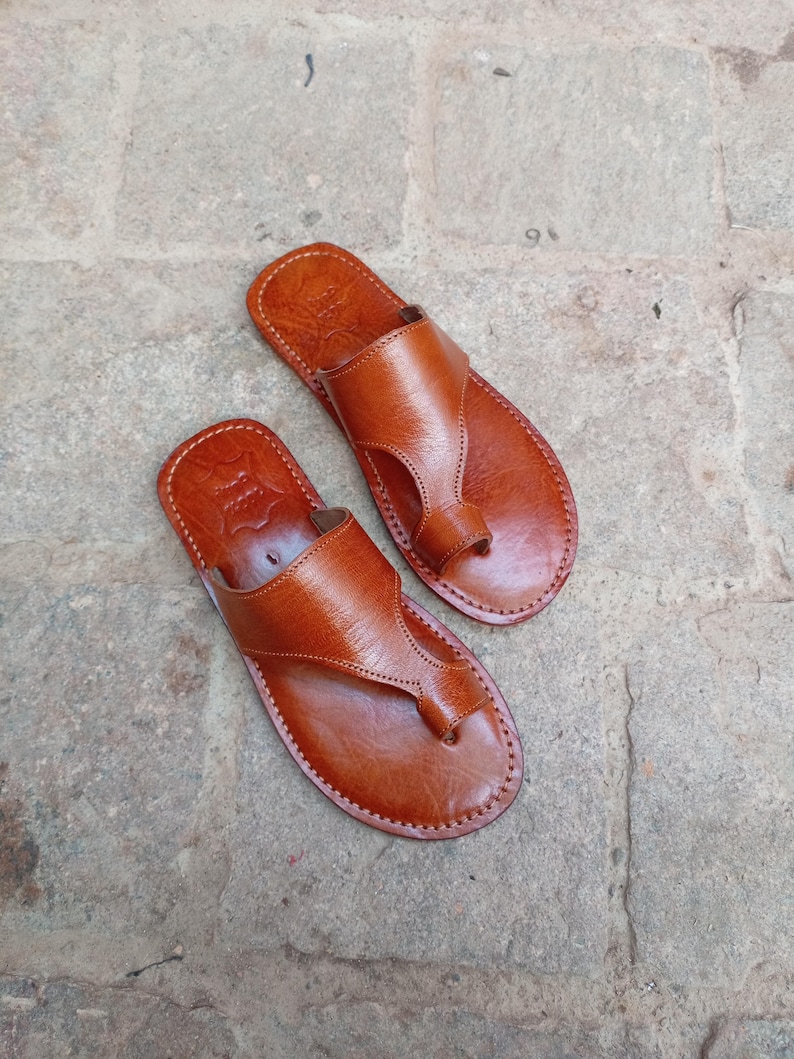May include: A pair of brown leather sandals with a thong-style design. The sandals have a smooth, polished appearance and are stitched around the edges. The sandals are designed for casual wear.