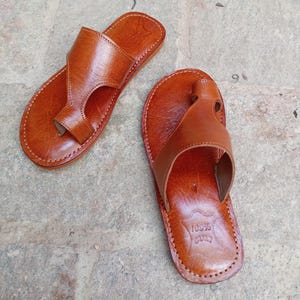 May include: A pair of brown leather sandals with a thong-style design. The sandals have a stitched sole and a strap that goes over the top of the foot. The leather has a natural, textured appearance. The sandals are likely handmade.
