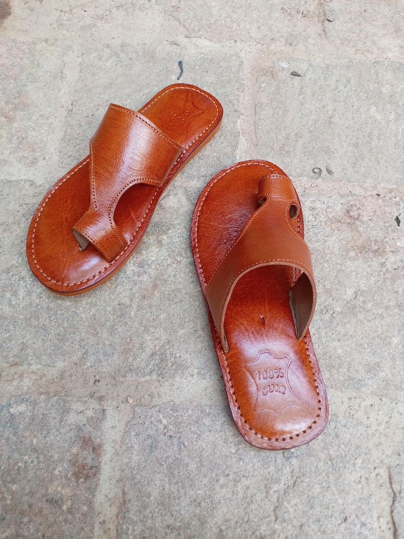 May include: A pair of brown leather sandals with a thong-style design. The sandals have a stitched sole and a strap that goes over the top of the foot. The leather has a natural, textured appearance. The sandals are likely handmade.