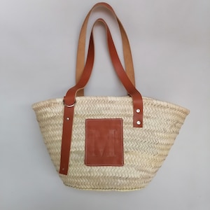 Op de afbeelding: Geweven strooien tote bag met bruine leren handvatten en accenten. De tas heeft een rechthoekige bruine leren patch met de letter "M" in reliëf op de voorkant. De handvatten zijn bevestigd met metalen ringen en klinknagels.