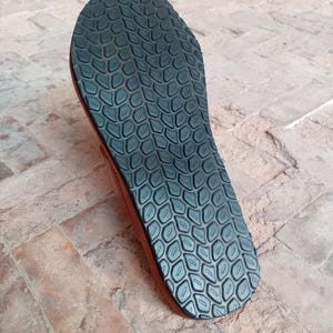 May include: Close-up of the sole of a brown sandal. The sole is black with a textured pattern. The sandal is made of leather and rubber, designed for comfort and durability. The sandal is likely for outdoor wear.