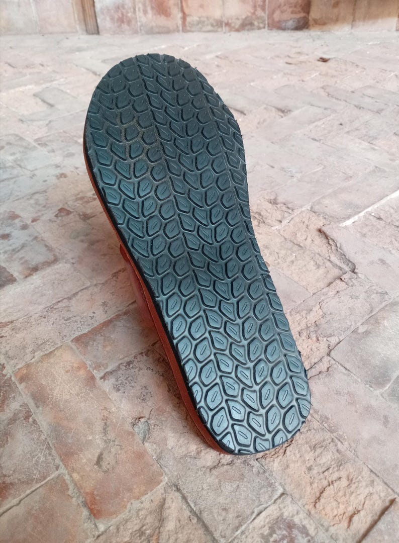 May include: Close-up of the sole of a brown sandal. The sole is black with a textured pattern. The sandal is made of leather and rubber, designed for comfort and durability. The sandal is likely for outdoor wear.
